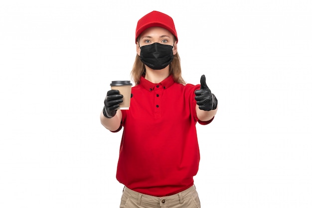 A front view female courier in red shirt black gloves mask and red cap holding cofffe cup showing like sign