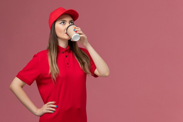 Front view female courier in red uniform and cape drinking coffee on the pink background service delivery uniform job worker