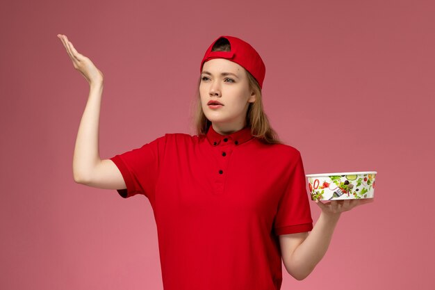 Front view female courier in red uniform and cape holding delivery bowl on light-pink wall, service uniform delivery worker job work