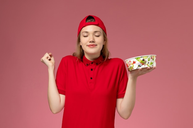 Front view female courier in red uniform and cape holding delivery bowl on light-pink wall, worker job service uniform delivery