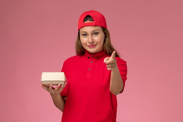 Front view female courier in red uniform and cape holding delivery food package on pink background uniform delivery service company job work worker