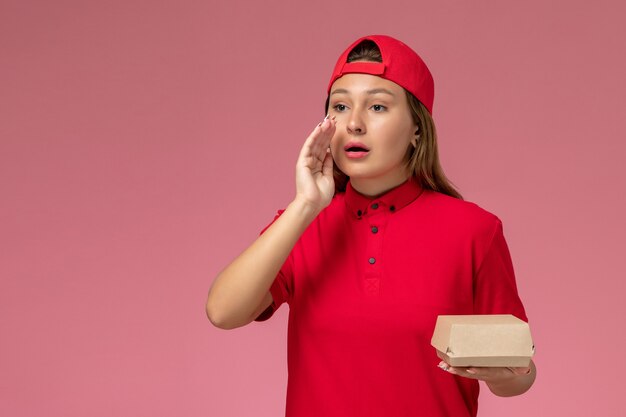 Front view female courier in red uniform and cape holding delivery food package on the pink wall, uniform delivery service company worker work girl