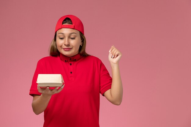 Front view female courier in red uniform and cape holding little delivery food package on light-pink desk uniform delivery service company