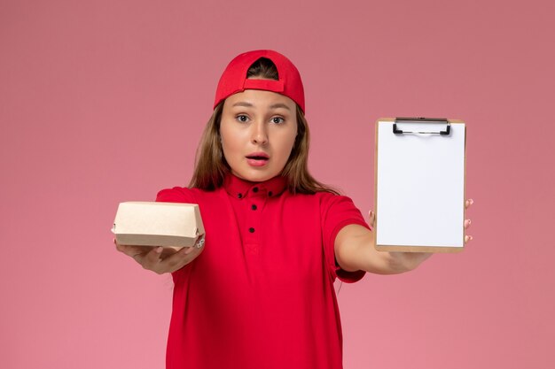 Front view female courier in red uniform and cape holding little delivery food package and notepad on the pink wall, uniform delivery service company worker