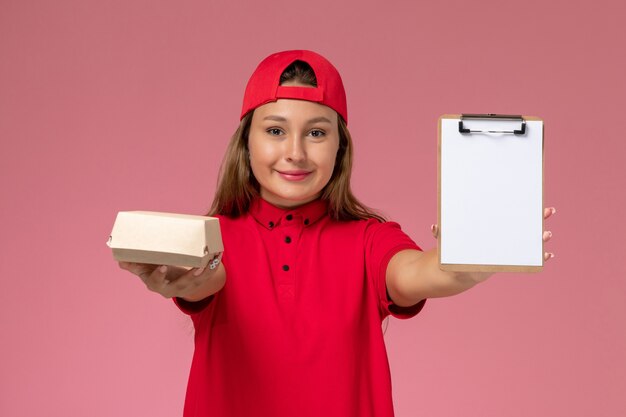 Front view female courier in red uniform and cape holding little delivery food package and notepad on the pink wall, uniform delivery service worker company