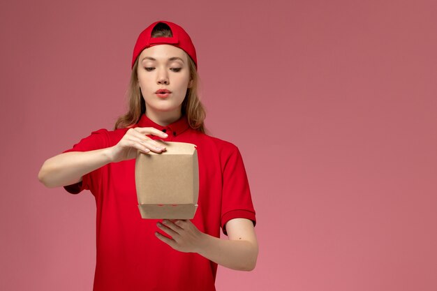 Front view female courier in red uniform and cape holding little delivery food package opening it on pink wall, worker delivery service company uniform