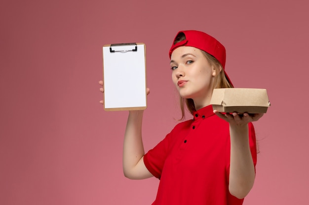 Free photo front view female courier in red uniform and cape holding little delivery food package with notepad on pink wall, delivery service uniform job worker work