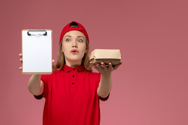 Front view female courier in red uniform and cape holding little delivery food package with notepad on pink wall, delivery service uniform job worker