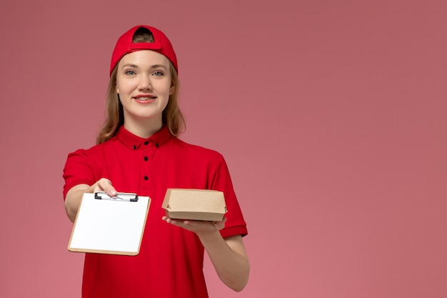 Free Photo front view female courier in red uniform and cape holding little delivery food package with notepad on the pink wall, service uniform delivery worker job