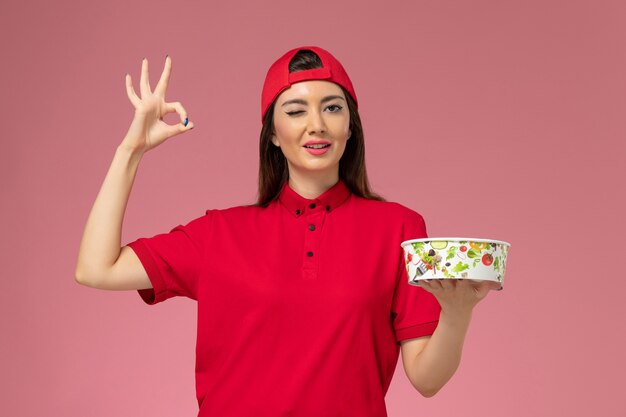 Front view female courier in red uniform cape with delivery bowl on her hands on light pink wall, uniform delivery employee job