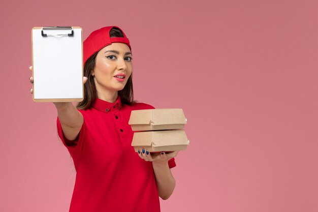 Front view female courier in red uniform cape with notepad and little delivery food packages on her hands on light-pink wall, job service delivery employee