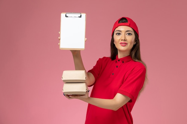 Free photo front view female courier in red uniform cape with notepad and little delivery food packages on her hands on light-pink wall, service delivery employee worker job