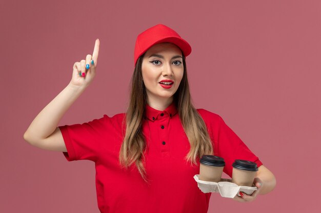 Front view female courier in red uniform holding brown delivery coffee cups on the pink background service delivery uniform job