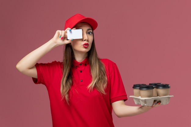 Front view female courier in red uniform holding delivery coffee cups and card on pink background service delivery company job uniform