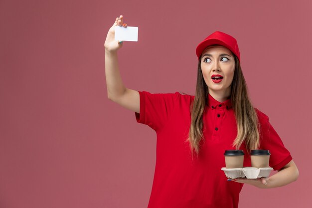 Front view female courier in red uniform holding delivery coffee cups and card on the pink background service job delivery uniform