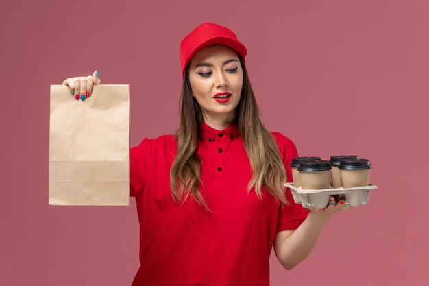 Front view female courier in red uniform holding delivery coffee cups with food package on the pink background service delivery job uniform