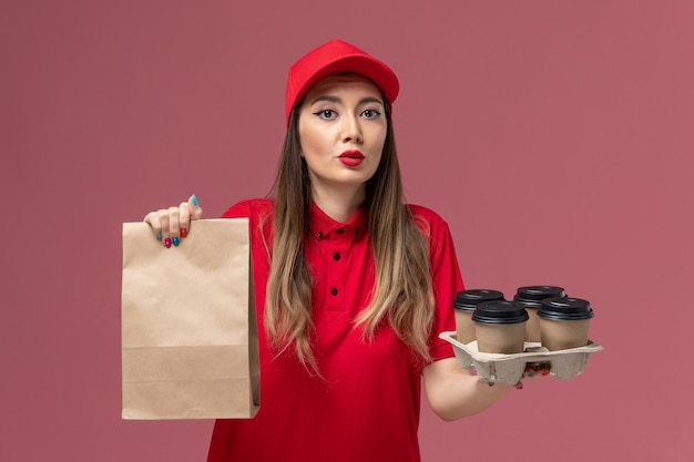 Front view female courier in red uniform holding delivery coffee cups with food package on pink background service delivery uniform