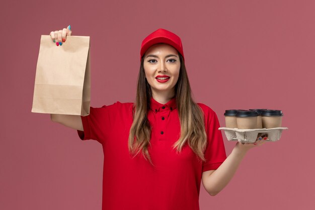 Front view female courier in red uniform holding delivery coffee cups with food package on the pink desk service delivery job uniform