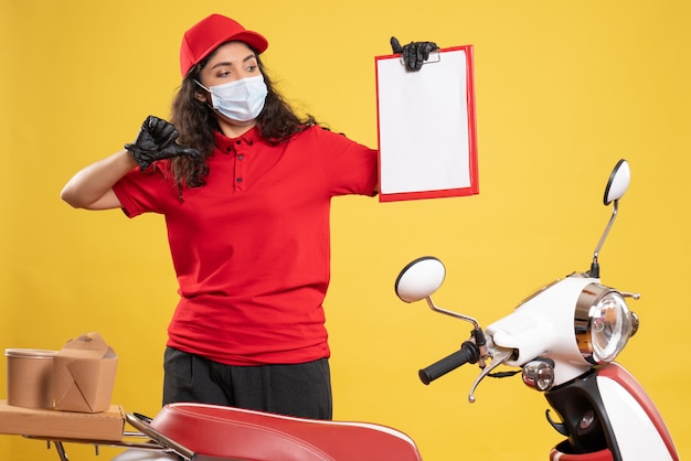 Front view female courier in red uniform holding file note on the yellow background delivery covid- service uniform worker pandemic job