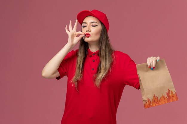 Front view female courier in red uniform holding paper food package showing tasty sign on pink background service job delivery uniform company