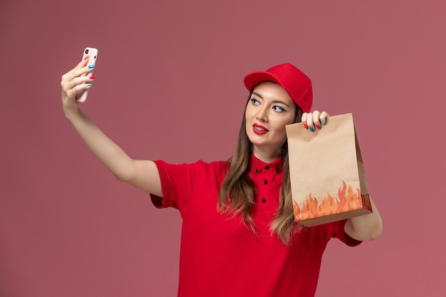 Free photo front view female courier in red uniform holding phone and food package taking photo on the pink background service delivery uniform job worker