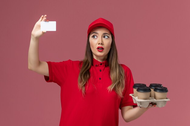 Front view female courier in red uniform holding white card on light pink background service job delivery uniform company