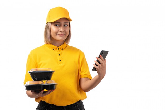 A front view female courier in yellow shirt yellow cap and black jeans holding bowls with food and smartphone smiling on white