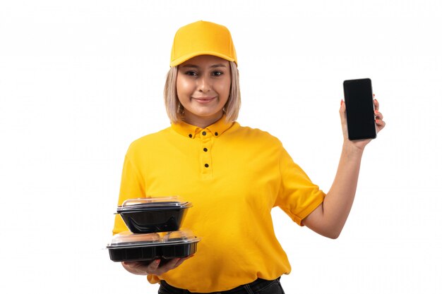 A front view female courier in yellow shirt yellow cap and black jeans holding bowls with food and smartphone smiling on white