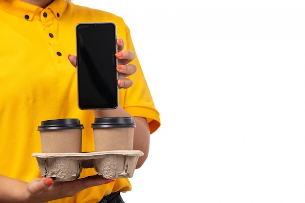 Free photo a front view female courier in yellow shirt yellow cap and black jeans holding coffee cups and smartphone on white