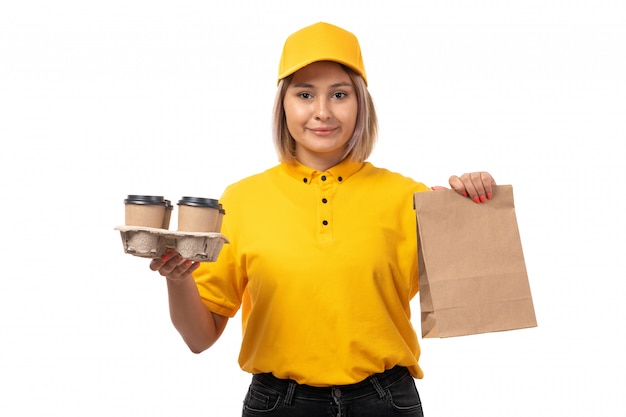 A front view female courier in yellow shirt yellow cap smiling holding coffee cups on white