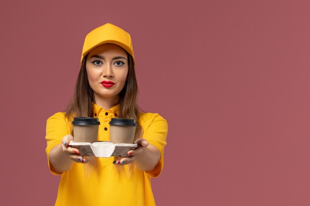 Front view of female courier in yellow uniform and cap holding brown delivery coffee cups on pink wall