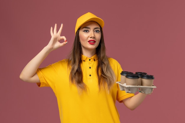 Front view of female courier in yellow uniform and cap holding brown delivery coffee cups on pink wall