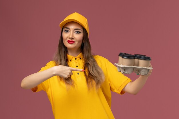 Front view of female courier in yellow uniform and cap holding brown delivery coffee cups on the pink wall