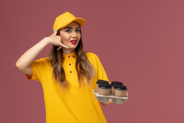 Front view of female courier in yellow uniform and cap holding brown delivery coffee cups on the pink wall