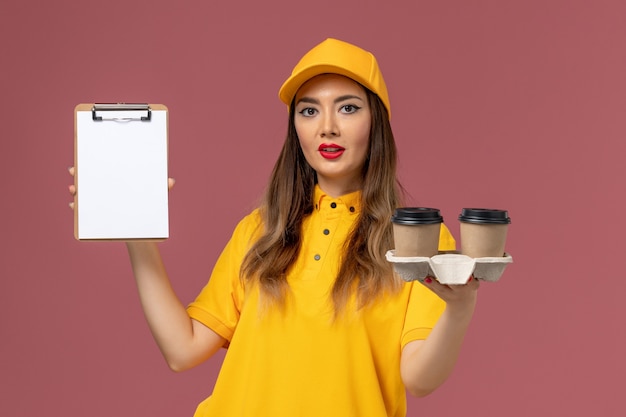 Front view of female courier in yellow uniform and cap holding delivery coffee cups and notepad on light pink wall