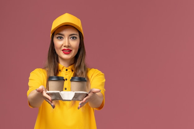 Front view of female courier in yellow uniform and cap holding delivery coffee cups on pink wall