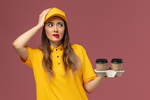 Front view of female courier in yellow uniform and cap holding delivery coffee cups on the pink wall