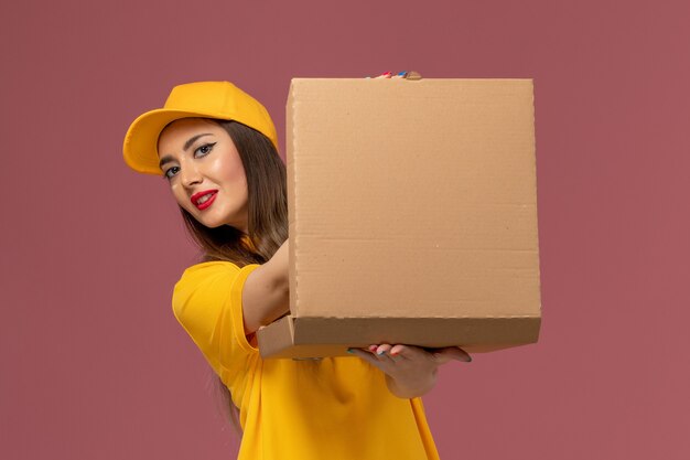 Free Photo front view of female courier in yellow uniform and cap holding food box on light pink wall