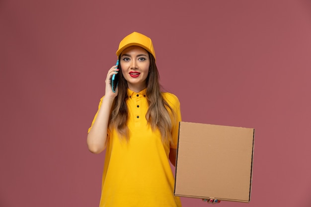 Free Photo front view of female courier in yellow uniform and cap holding food box and talking on the phone on light pink wall