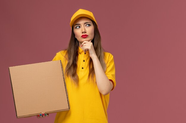 Front view of female courier in yellow uniform and cap holding food box thinking on the light-pink wall