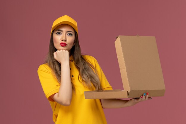 Front view of female courier in yellow uniform and cap holding open food box on the light pink wall