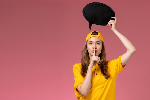 Front view female courier in yellow uniform and cape holding black sign on the pink wall company service delivery uniform job work