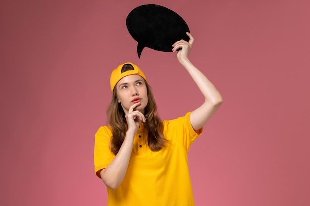 Front view female courier in yellow uniform and cape holding black sign and thinking on pink wall service delivery uniform