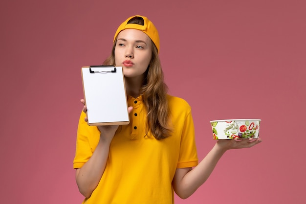 Front view female courier in yellow uniform and cape holding delivery bowl and notepad thinking on the light pink wall service delivery uniform company job