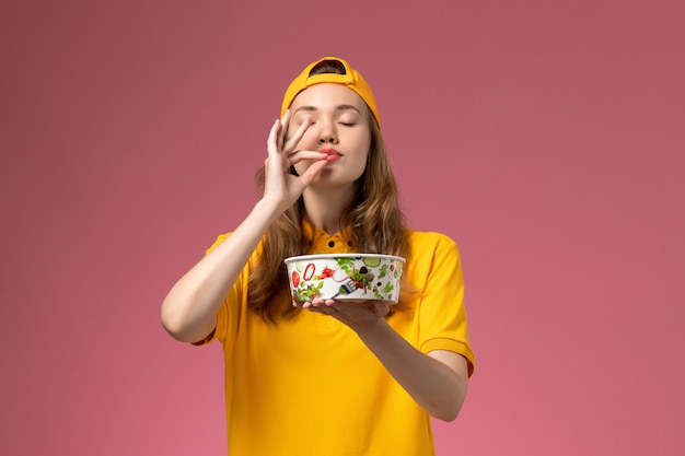 Front view female courier in yellow uniform and cape holding delivery bowl on the pink wall service delivery uniform company job work