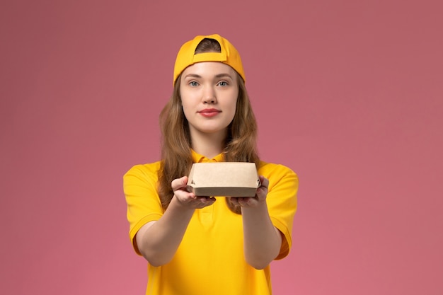 Front view female courier in yellow uniform and cape holding delivery food package on pink wall service delivery uniform company job worker girl work