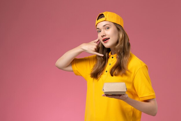 Front view female courier in yellow uniform and cape holding delivery food package on pink wall service delivery uniform worker girl work