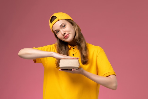 Front view female courier in yellow uniform and cape holding empty little delivery food package on the light pink wall service delivery uniform