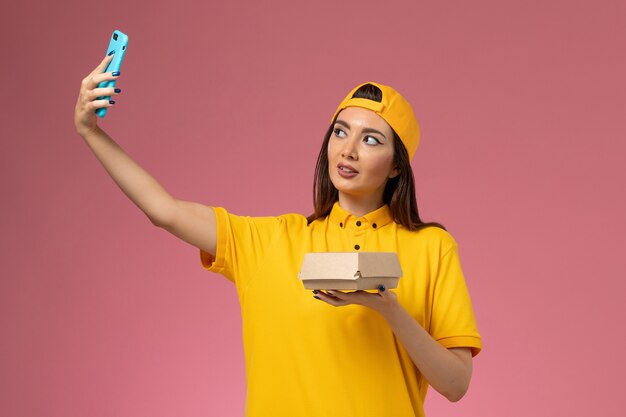 Front view female courier in yellow uniform and cape holding food package and taking photo on light pink wall company service uniform delivery