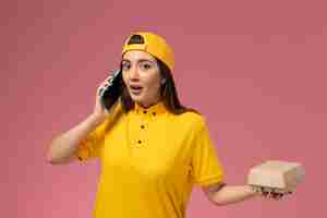 Free photo front view female courier in yellow uniform and cape holding food package and talking on the phone on the light-pink wall company service uniform delivery work